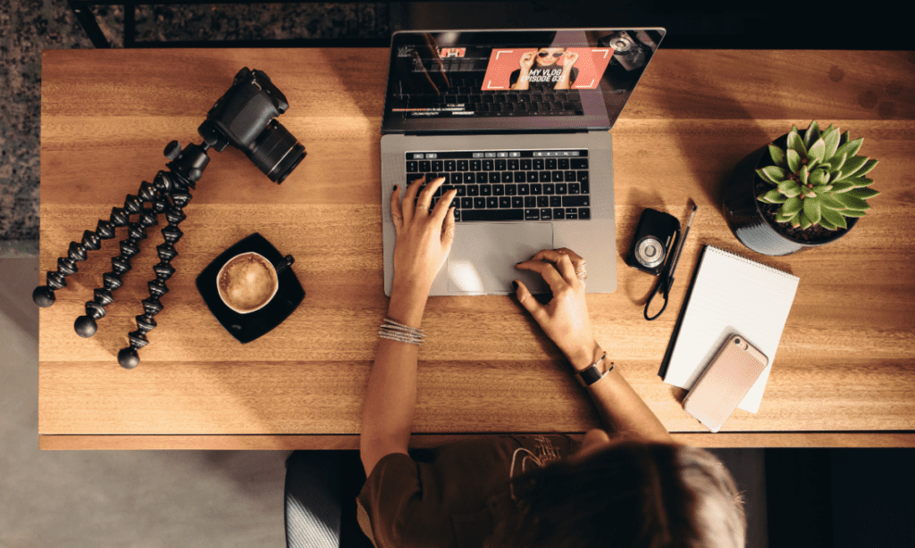A content creator sits in front of her computer with camera gear on the desk, exemplifying the benefit of having a sustainable social media strategy for driving sales through ads. 
